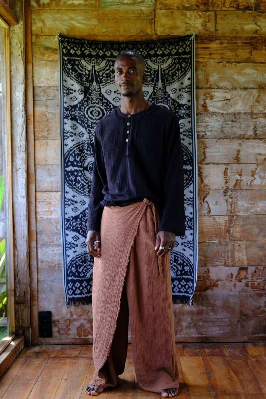 Model wearing Nahara Pants Brown - Men with a black shirt, showcasing relaxed summer fashion in a tropical setting.
