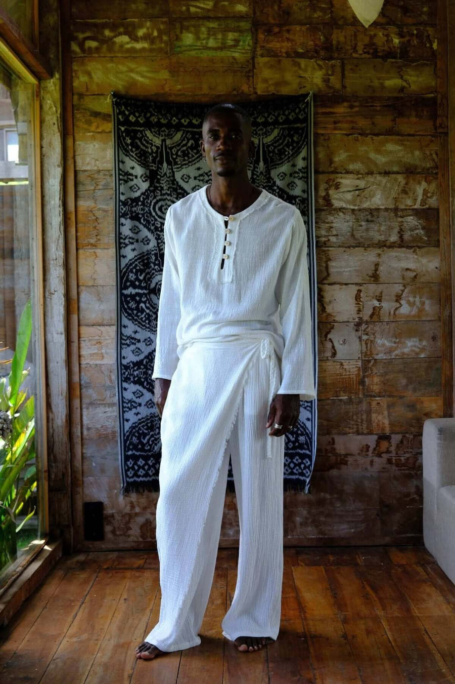 Nahara Pants White - Men worn by model in a relaxed setting with wooden decor and tropical elements.